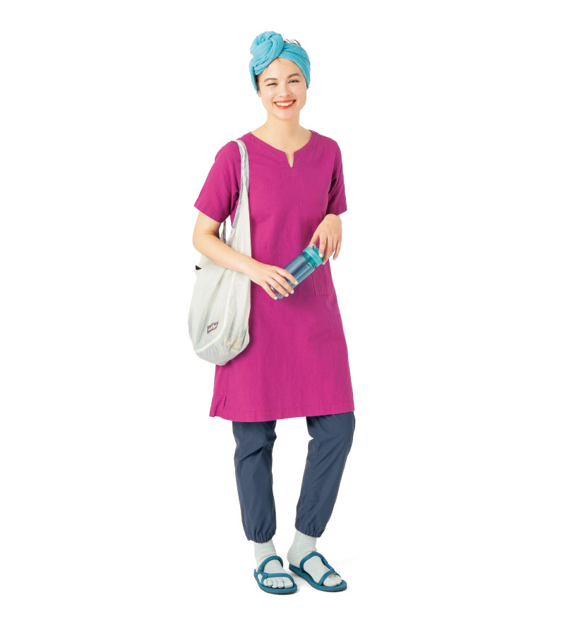 紅紫色の半袖ワンピースの下にネイビーのパンツを着用した女性モデル。水色のタオルマフラーを頭に巻き、ライトグレーのショルダーバッグを肩からさげ、青いサンダルを履いている