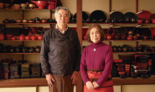 漆器が並ぶ棚の前に藤八屋の代表と女将が立っている